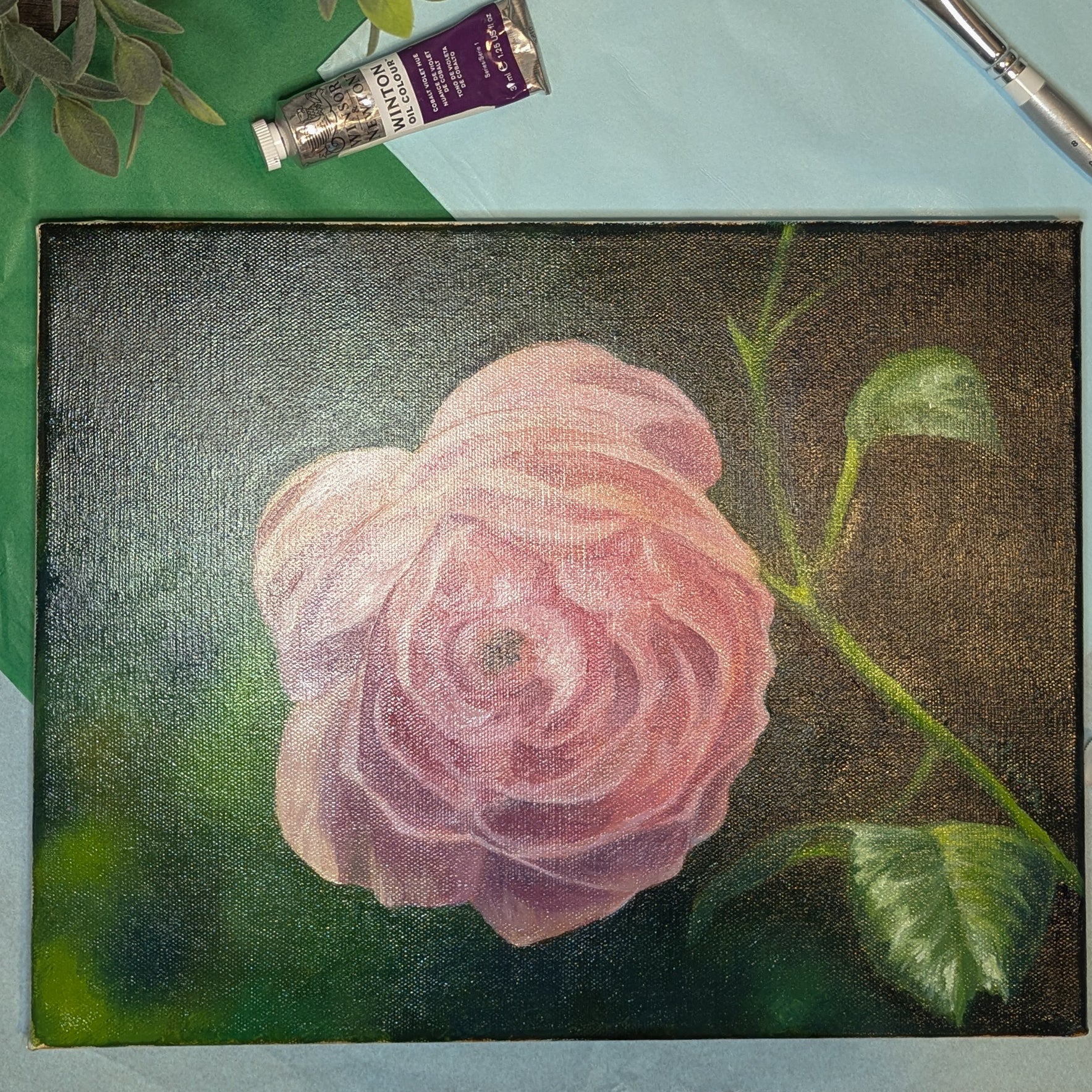 Painting of a pink rose on a textured surface with a tube of paint and brush in the background.