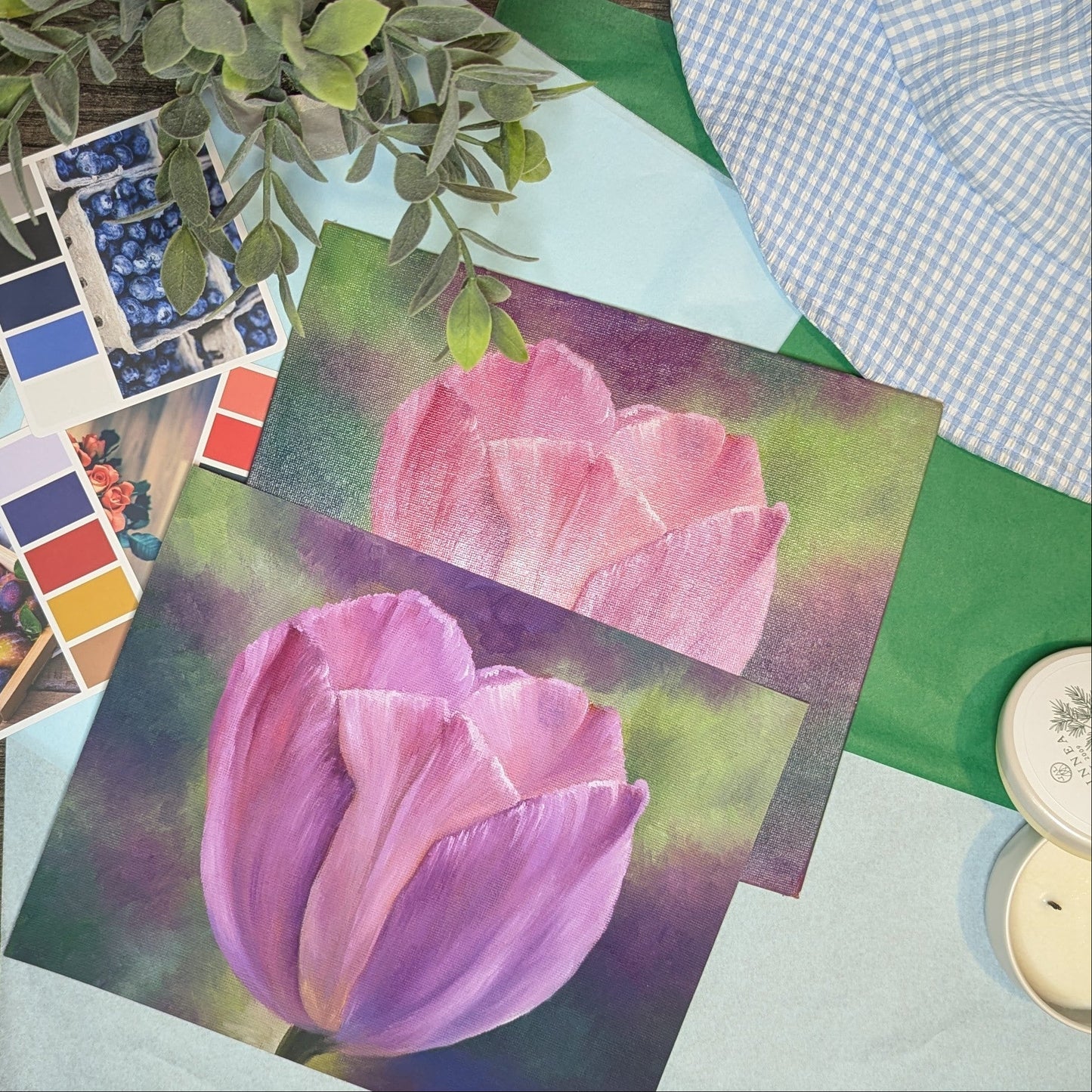 Original oil painting of a tulip next to the print of the painting. Color swatches, flowers, and a plant on a table with a checkered cloth in the background.