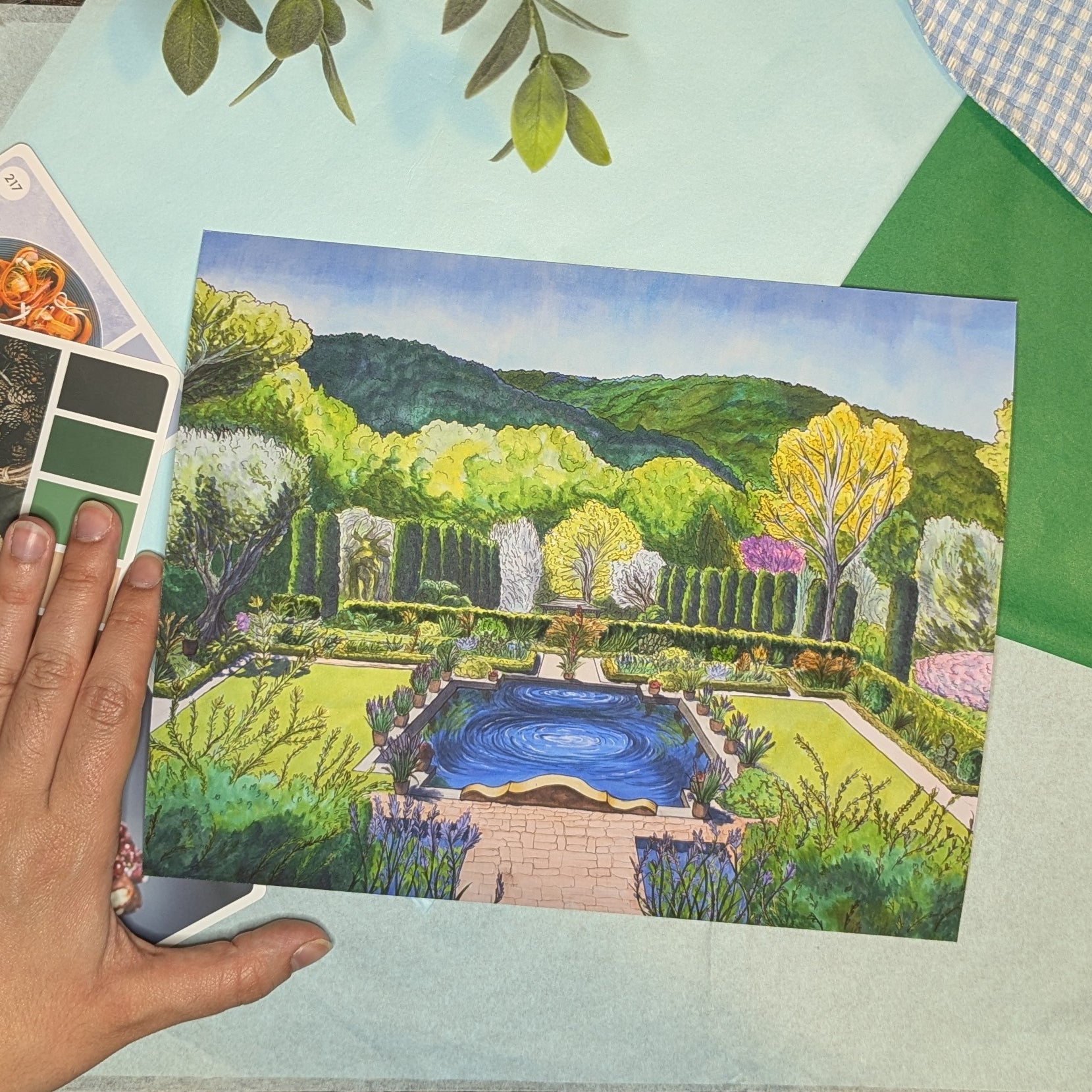 Hand holding a painting of a garden with a pool on a green surface