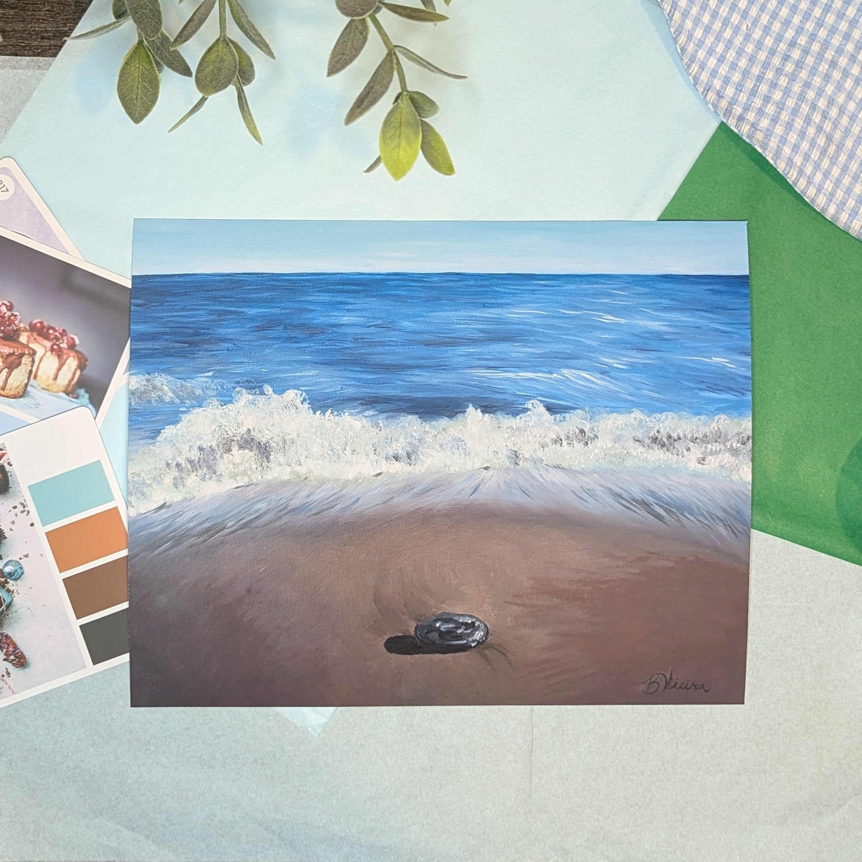 Painting of a beach scene with waves and a rock, placed on a surface with art supplies.