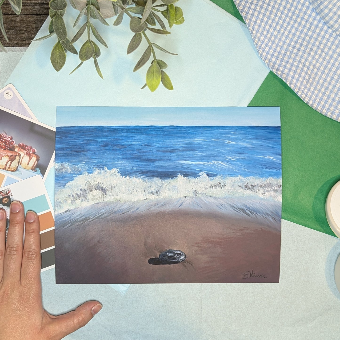 Hand holding a beach scene painting on a colorful surface with greenery.